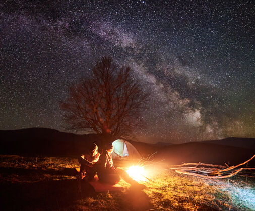 银河夜间露营在星空下的营火旁休息的徒步旅行者帐篷全景放松