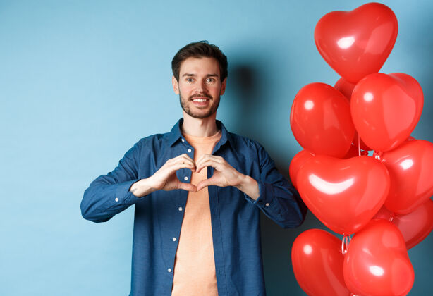 温柔情人节快乐恋爱中的男朋友向爱人展示心形 微笑着 站在蓝色背景下的红气球旁心情人情感