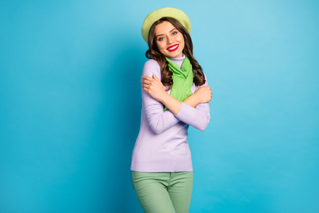 情感迷人女士拥抱自己的照片享受和谐牙齿微笑温暖的衣服穿绿色贝雷帽紫色套头衫围巾裤子孤立的蓝色墙壁自我乐趣时尚