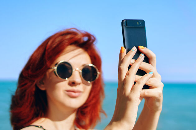 移动夏季自拍美女在蓝天碧海的映衬下自拍戴着墨镜的红发女子在智能手机摄像头上自拍使异国情调美丽
