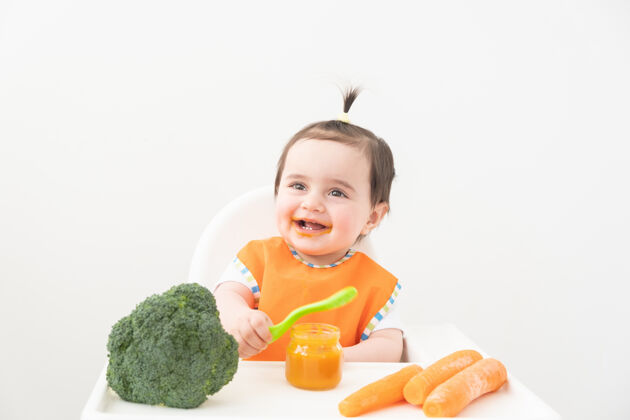 保健穿着橙色围嘴的女婴坐在儿童椅上吃白色背景的蔬菜泥餐父母母亲