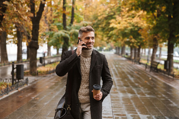 手机一张30多岁的时尚男士穿着暖和的衣服 在户外穿过秋日公园 并使用手机的照片成人白种人男士