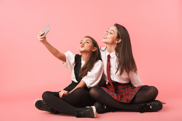 积极穿着校服的有趣女孩坐在隔着红墙的地板上用手机自拍的照片请白种人奥本