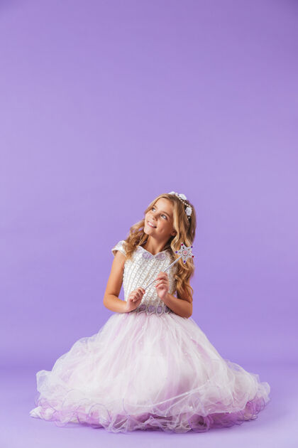 欢乐穿着公主裙的漂亮小女孩孤零零地坐在紫罗兰色的墙上 手里拿着魔杖快乐五颜六色小