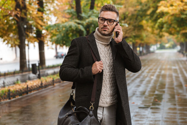 成人一张30多岁的严肃男人穿着暖和的衣服在户外穿过秋天公园的照片 正在讲手机交谈享受夹克