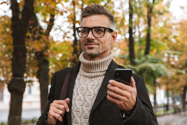 站立30多岁的留胡子男人穿着暖和的衣服 在户外穿过秋天公园 用手机拍照吸引力帅气户外