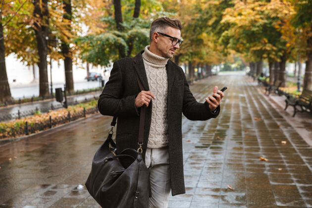 胡须30多岁的时尚小伙穿着暖和的衣服在户外穿过秋天公园 用手机拍照夹克阳刚散步
