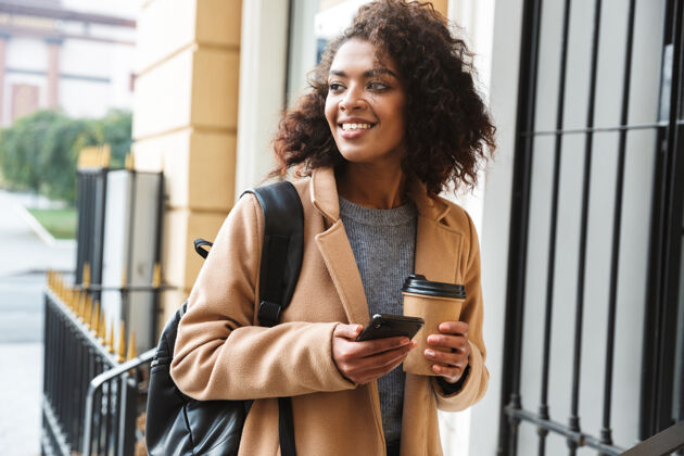 城市快乐的非洲年轻女子穿着外套走在户外 拿着外卖咖啡杯 用手机休闲微笑电话