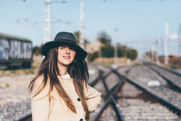 时髦在废弃的铁轨上穿着米色雨衣戴着帽子的快乐女人轨道经典女性化