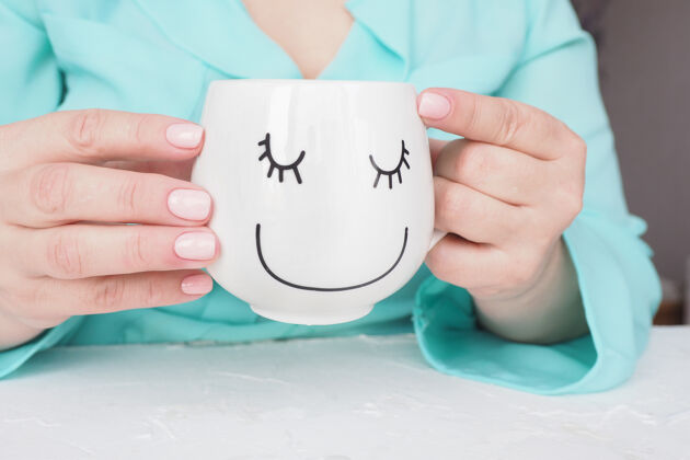 手美丽的美甲和柔软的粉红色指甲油的女性手拿着一杯咖啡或茶 一张有趣的画脸美甲咖啡衬衫