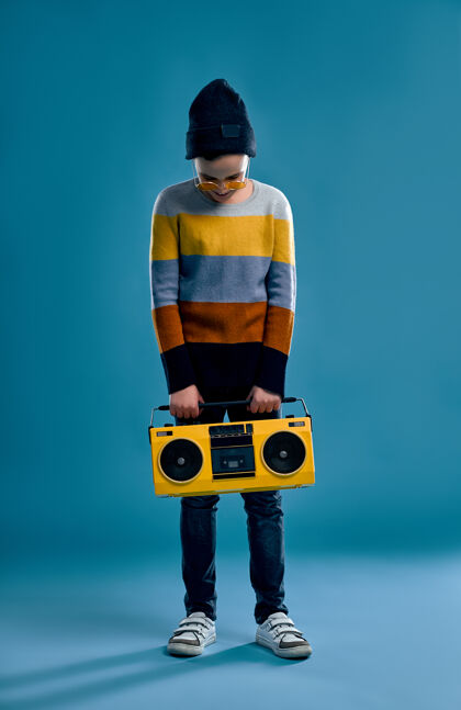 帅气一个穿着条纹毛衣 戴着帽子 戴着黄色眼镜的时髦男孩手里拿着一台复古的录音机 一台蓝色的录音机磁带唱片播放器
