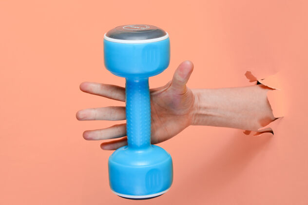 主动女性的手拿着一个蓝色的塑料哑铃穿过撕破的粉色纸简约运动理念质量健身房撕裂