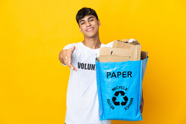 垃圾一名男子拿着一个装满废纸的回收袋在孤立的黄色背景上进行回收 握手表示成交不错合作年轻人环境