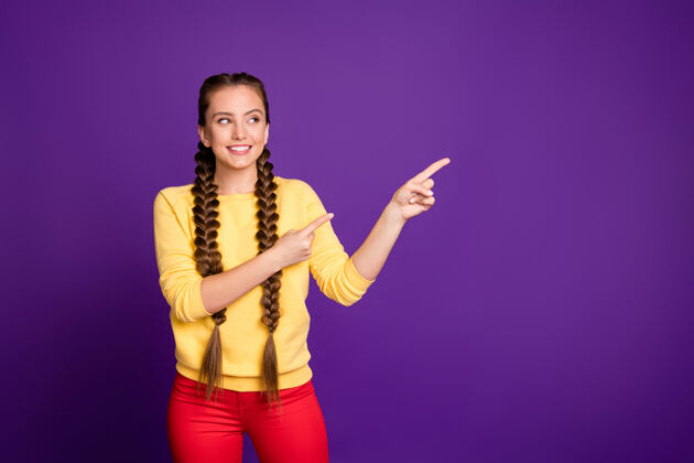 促销搞笑女士长辫子发式表示手指空位穿休闲黄色套头衫红色长裤隔离紫色墙壁推荐青少年裤子