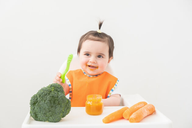 胡萝卜穿着橙色围嘴的女婴坐在儿童椅上吃着白色的蔬菜泥母亲食物营养
