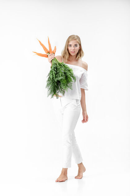 成人一个穿着白衬衫的金发美女 手里拿着一根新鲜的胡萝卜 上面有绿叶背景.健康还有节食食物自然生活方式