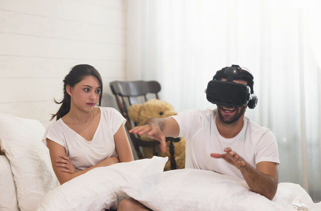 酒店女人躺在床上 她的丈夫留着黑色短发 留着3天的胡子 戴着3d眼镜用他的vr头戴式游戏视频 玩得很开心游戏玩妻子