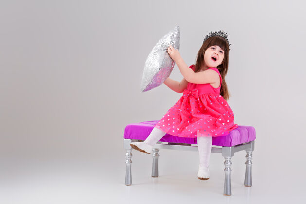 灰色穿着粉色公主裙 戴着王冠的漂亮的小黑发女孩坐在粉色的椅子上玩枕头可爱宝贝粉色微笑美丽