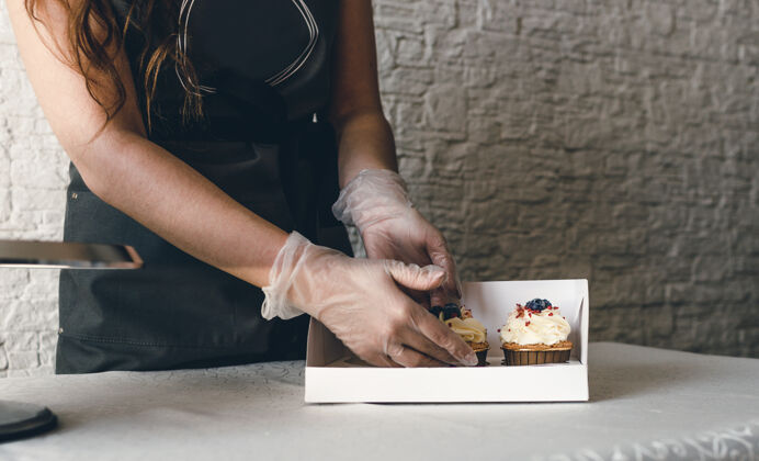 纸杯蛋糕一个穿着灰色围裙的女孩在一个礼品盒里包上奶油纸杯蛋糕 把订单送到餐厅客户.烘焙在家里食物包装烹饪