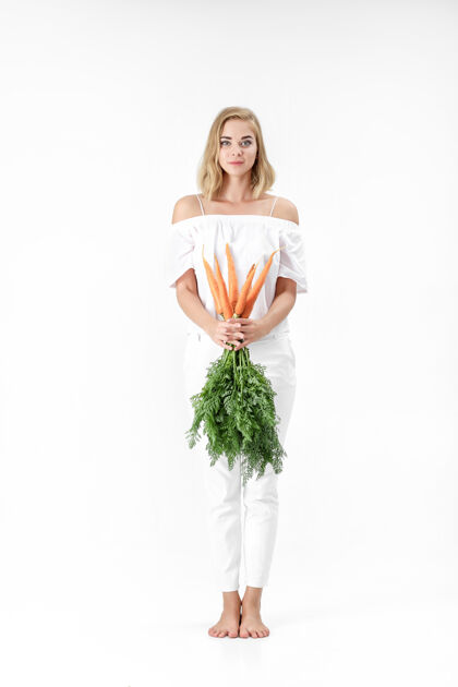 欢呼一个穿着白衬衫的金发美女 手里拿着一根新鲜的胡萝卜 上面有绿叶背景.健康还有节食白色成人素食