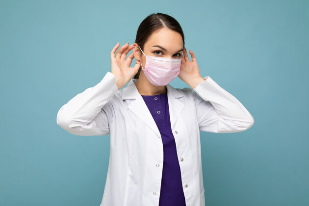 吸引身穿白大褂 戴着医用口罩的年轻女医生孤零零地站在蓝色上空的照片面罩保健黑发