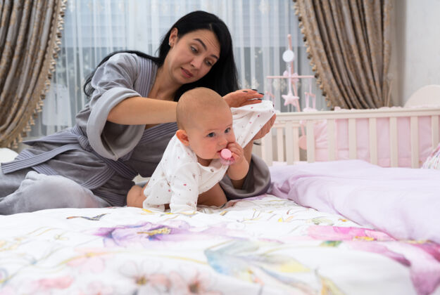 照顾一位沮丧的母亲穿着晨衣 在家里给婴儿穿衣 房间里有婴儿床可爱温柔托儿所