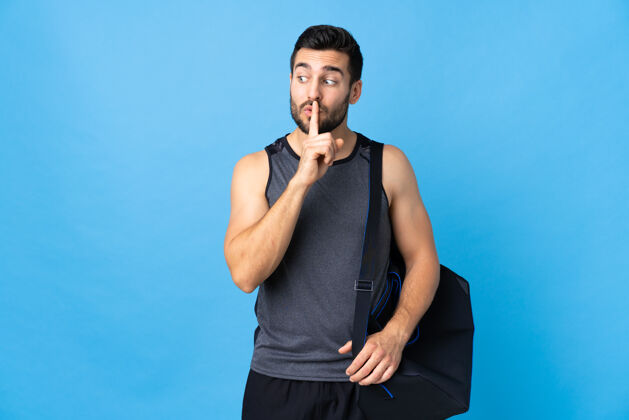 运动员在蓝色背景上 一个拿着运动包的年轻运动男表现出一种沉默的手势 把手指放进嘴里年轻安静护理