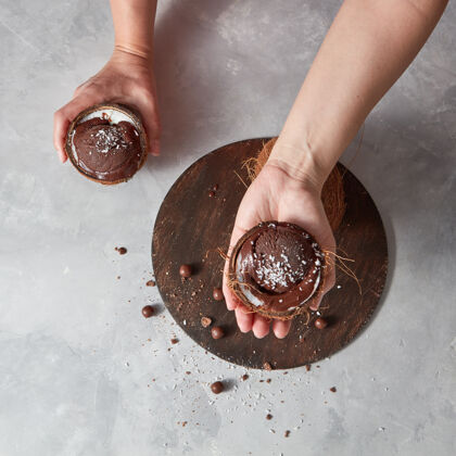 新鲜在一张灰色桌子上 一个椰子壳里的自制巧克力甜点 女人的手举在圆板上 桌上放着一个小蛋糕文本.夏季甜点贝壳排毒饮食融化