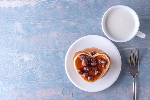 孩子自制的心形薄饼配浆果酱 白色牛奶杯和叉子放在蓝色的木桌上浆果浪漫情人节