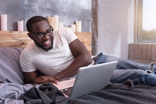 快乐快乐工作很高兴年轻的非洲男人戴着眼镜 躺在床上使用笔记本电脑男人室内帅气