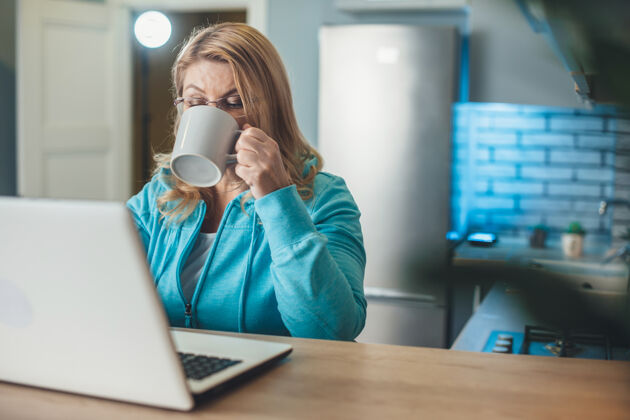女性忙碌的金发老太太正在厨房的笔记本电脑旁喝茶坐着认真远程