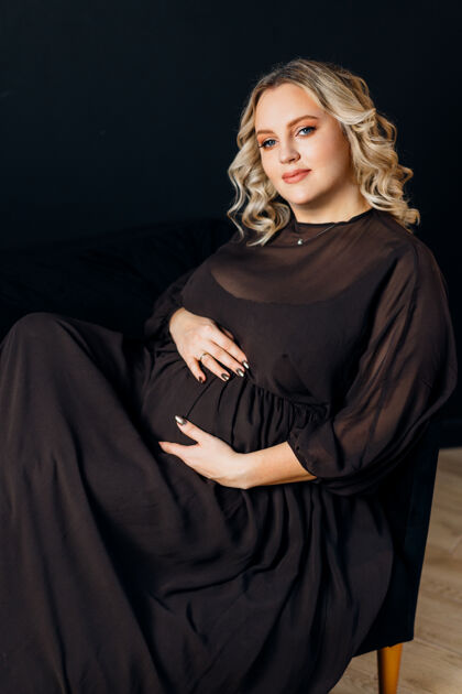 护理孕妇穿着优雅的黑色连衣裙在室内摄影棚摆姿势黑色墙壁背景金发白人中年女性怀孕6个月女人腹部新生活