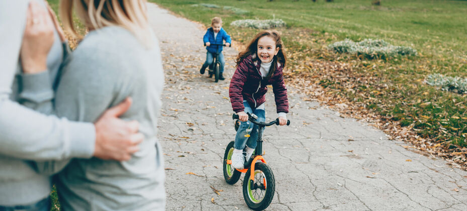 女性可爱的小女孩和小弟弟在公园里骑自行车 而父母正在看着他们夫妇乐趣看