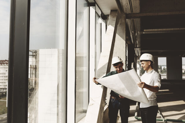 商业自信的业主微笑着拿着窗户旁边的建筑平面图和他的建筑师谈论着正在施工的建筑建筑工作执行