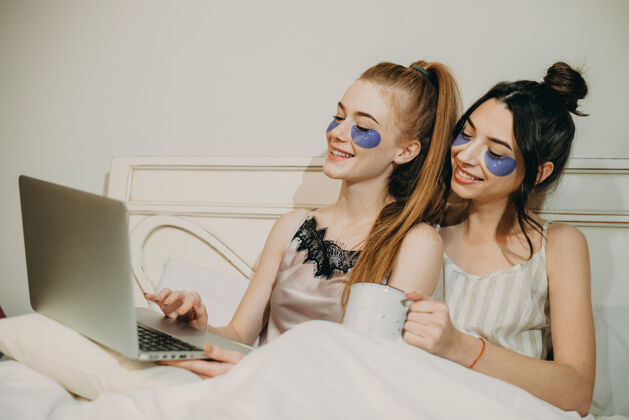 睡觉时间两个可爱的妹妹坐在床上微笑着喝着咖啡 看着一个眼睛上有补丁的笔记本电脑屏幕美丽女人皮肤护理