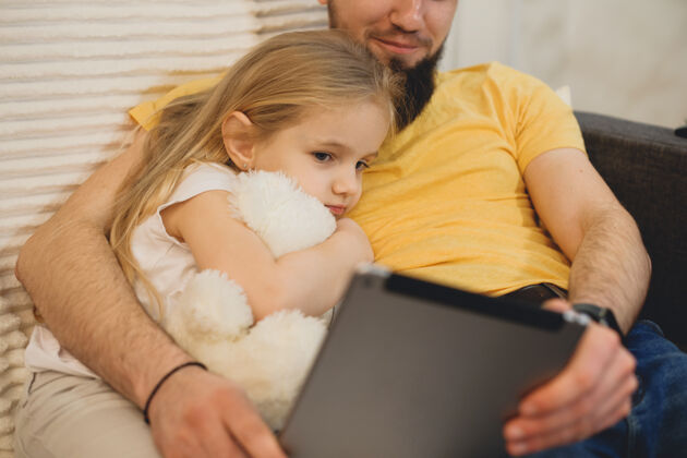 小一个可爱的小女孩靠在她父亲的胸前 抱着一只白色的泰迪熊 看着她父亲的平板电脑兄弟自由空间男人