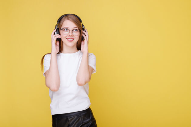 甜蜜戴眼镜的年轻女孩在黄色的墙上听音乐 微笑着音乐时尚配件