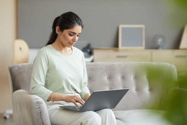 电脑坐在沙发上用笔记本电脑打字的年轻女子她在家上网工作看快乐家庭生活