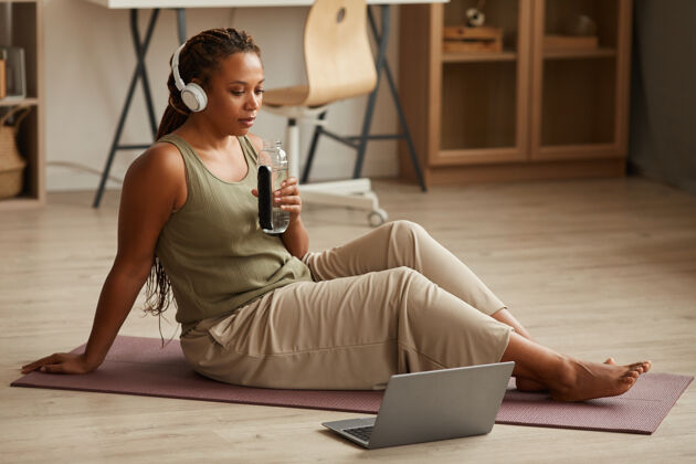 耳机戴着耳机的年轻女子坐在运动垫上喝水 在家里用笔记本电脑在线观看体育锻炼瑜伽通讯笔记本电脑
