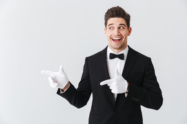 手势一位身穿燕尾服 戴着手套的帅哥侍者的肖像 孤立地站在白色的墙上 用手指着复印空间餐厅服务人
