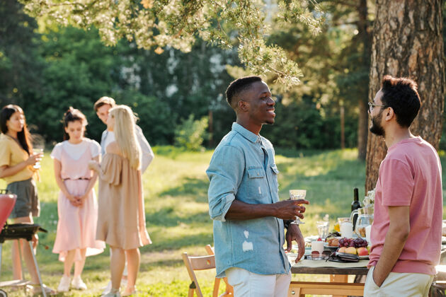 欢呼两个穿着休闲服的快乐的年轻人在户外晚餐后站在餐桌旁讨论最新的足球比赛一群人烧烤色彩形象