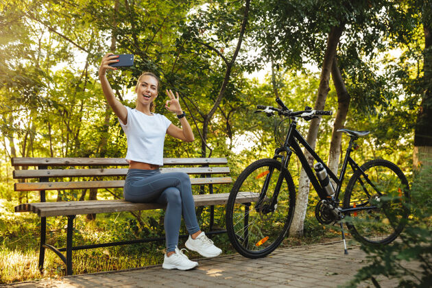 成人可爱的女人骑着自行车坐在公园的长椅上 用手机自拍阳光运动员休息