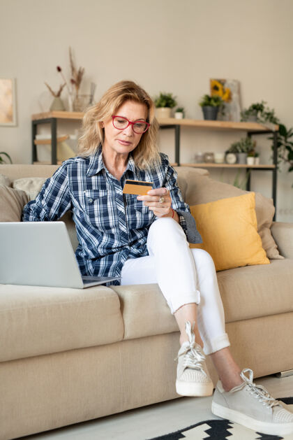 购买金发熟女坐在客厅笔记本电脑前的沙发上看信用卡 准备支付网上商品房子休闲服客厅