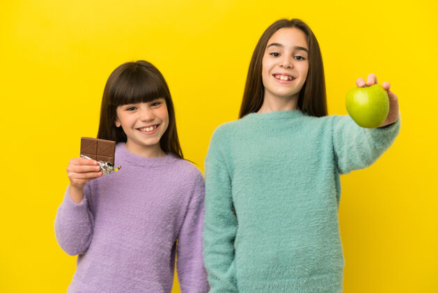 巧克力黄色背景下孤立的小姐妹们一手拿着巧克力片 一手拿着苹果乐趣拉美裔人