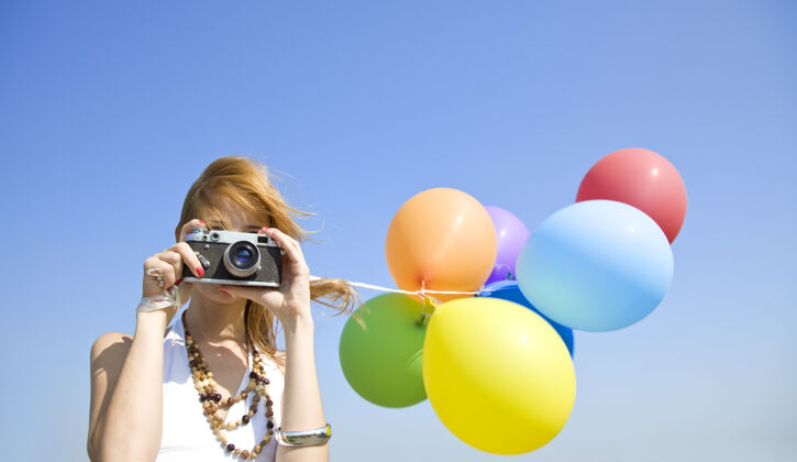 散步红发女孩带着彩色气球和照相机在蓝天上自由蓝色风