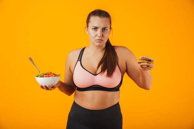 护理一个心烦意乱的超重健身妇女穿着运动服 孤立地站在黄色的墙上 手里拿着盛着沙拉和汉堡的碗不健康减肥健身