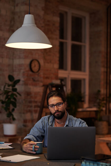 技术留着胡子的商人一边喝咖啡 一边看着笔记本电脑的显示器 一边在办公室的桌子旁工作长相计算机眼镜