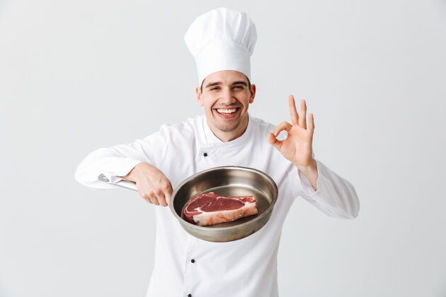 手开朗的厨师身着制服 展示平底锅和生牛排隔着白墙 展示ok餐馆厨房薯条