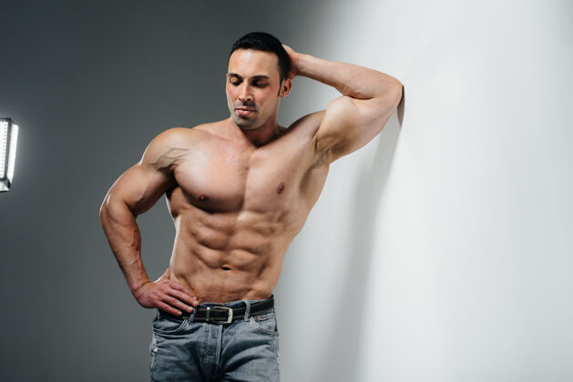 男子气概一个年轻的健美运动员赤裸上身穿着牛仔裤站在墙边运动身体力量近墙