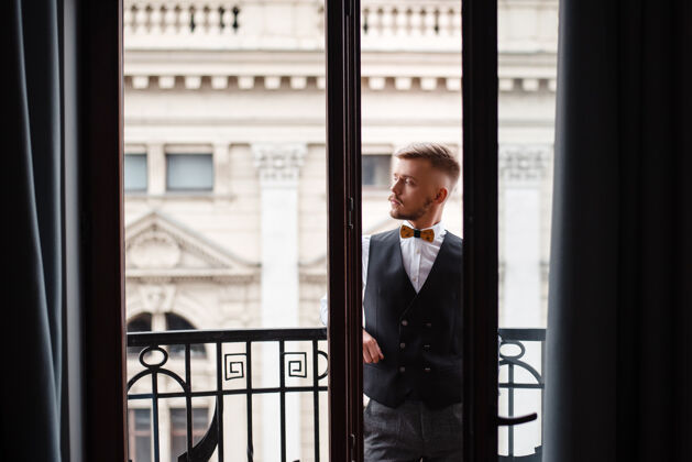 浪漫自信满脸胡须的新郎穿着时尚的婚纱站在酒店阳台上 一边看一边婚礼阳台时尚